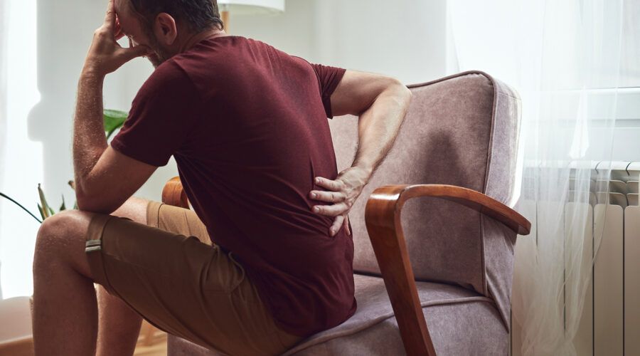 Mężczyzna siedzący na krześle trzyma się za plecy w geście oznaczającym ból kręgosłupa